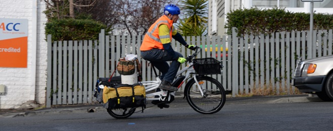 man wearing high-vis top and blue helmet riding white bicycle with bags on either side of the rear carrier and one bucket on top.