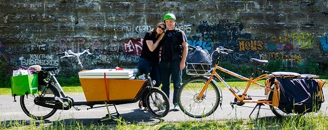 Two cargo bikes stand front wheel to front wheel with a man and woman standing behind them and a graffittied concrete wall in the background.