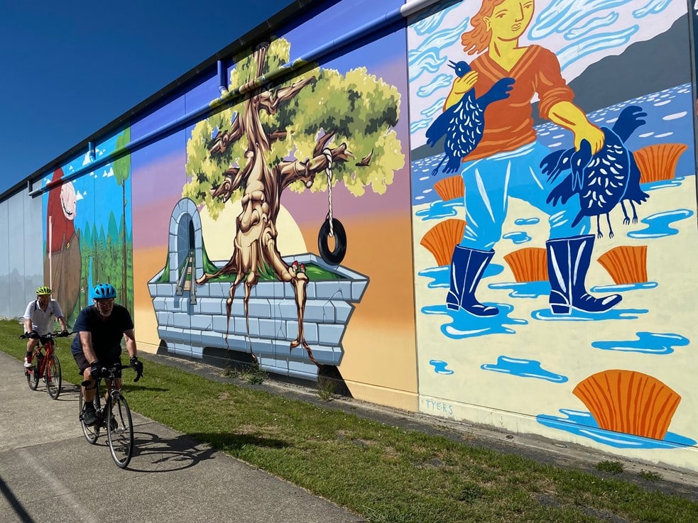 Two men ride past a triptych of three large murals on the back of a building along the Intercity Cycleway.