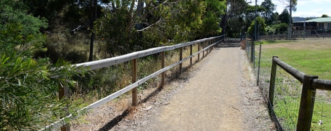Gravel path with wooden railings and bush on the left and grass on the right leads to a road junction.