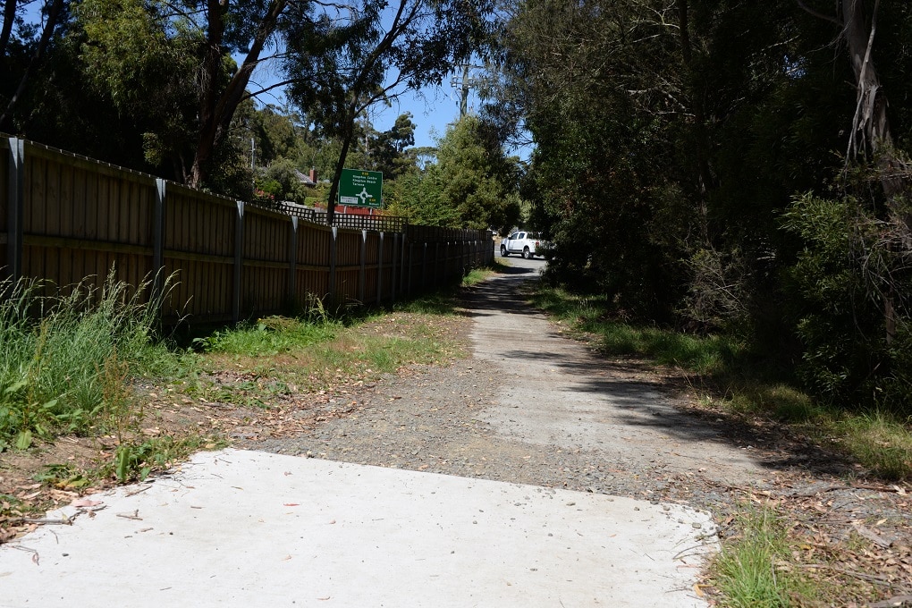 A wide concrete path ends and a narrower gravel trail leads to a road intersection, with a white car driving past.