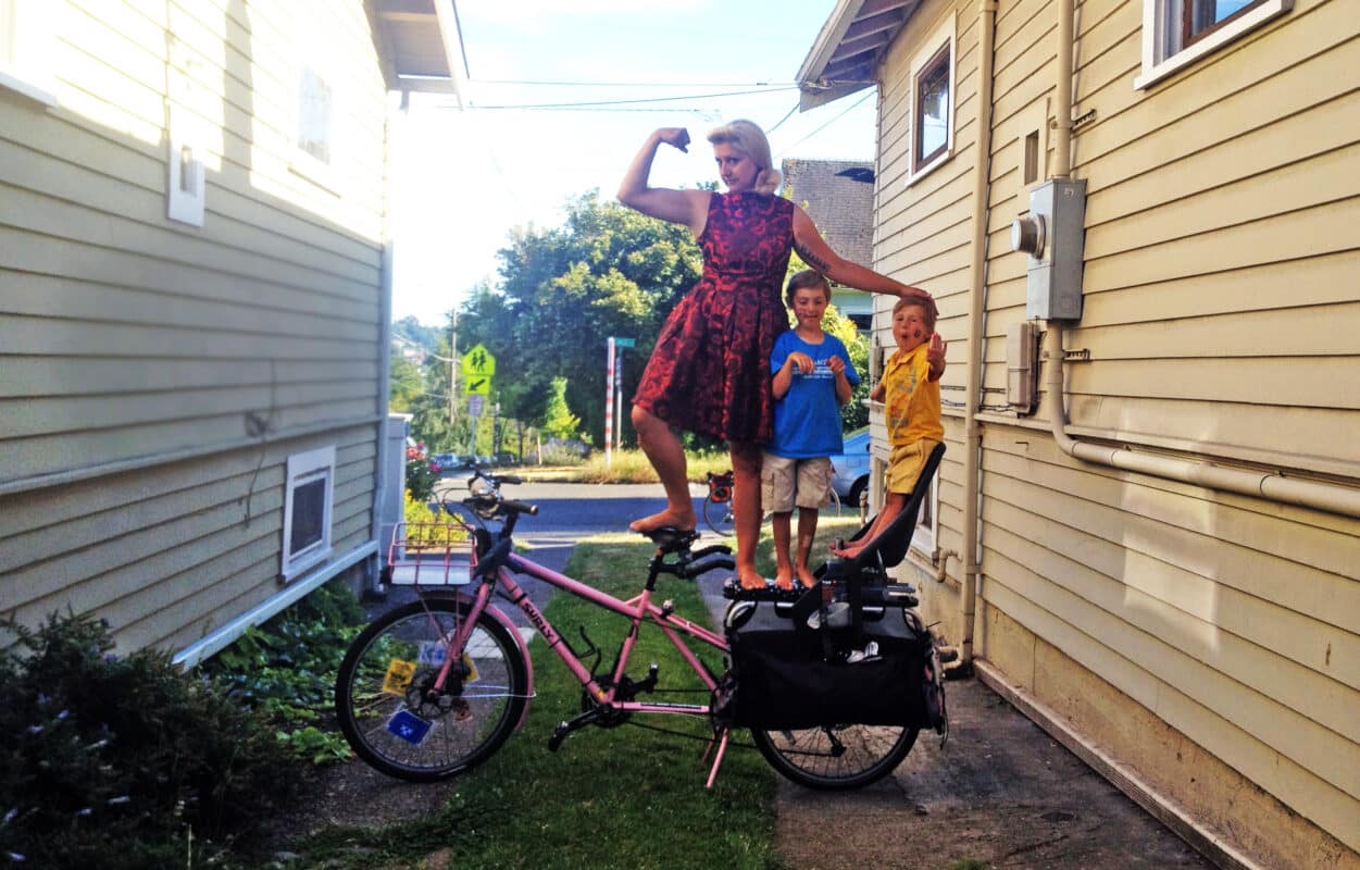 Woman in red and black 50s style dress stands on the seat and pack rack of her pink long-tail cargo bike with two young boys also standing on the pack rack, flanked by two weatherboard houses.