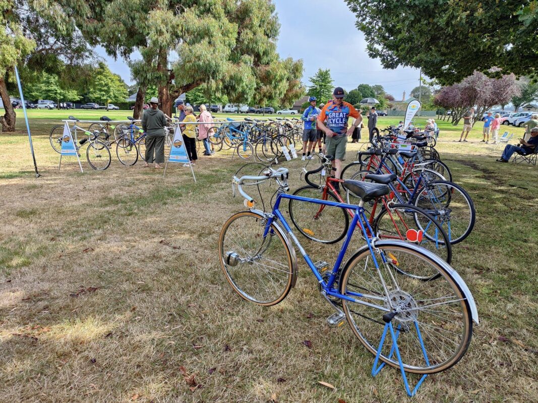 A bright blue old fashioned road bike sits at the end of a row of older style bikes, opposite another row of bikes while people mill around on the grass looking at the bikes. 