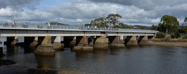 Image of an old bridge in a river with large concrete pylons and people on bikes lined up along the edge of the railing.