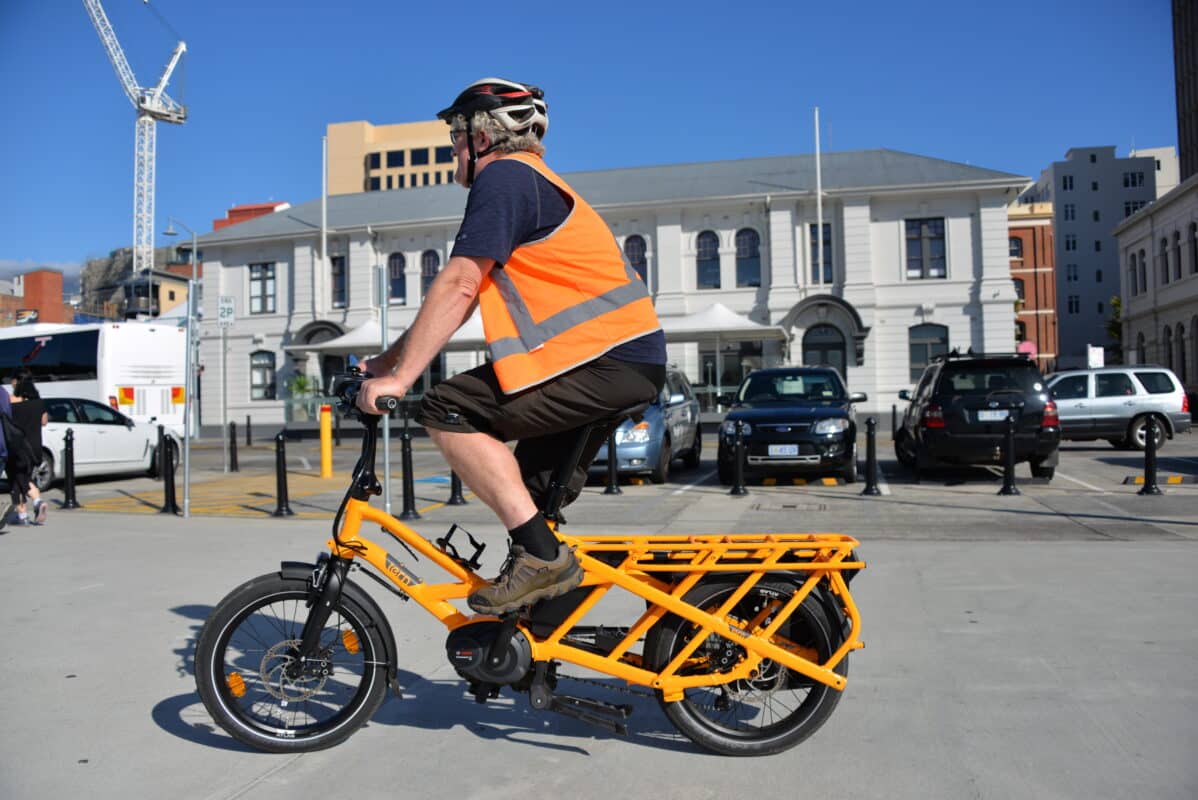 A man wearing an orange high vis vest and dark shorts and tshirt rides a bright orange Tern ebike along the Hobart waterfront path, with colonial buildings in the background. 