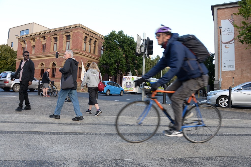 Slight blurred man rides past the lens in the foreground with people walking in different directions behind him and a man who is counting bikes stands still amidst the crowd.