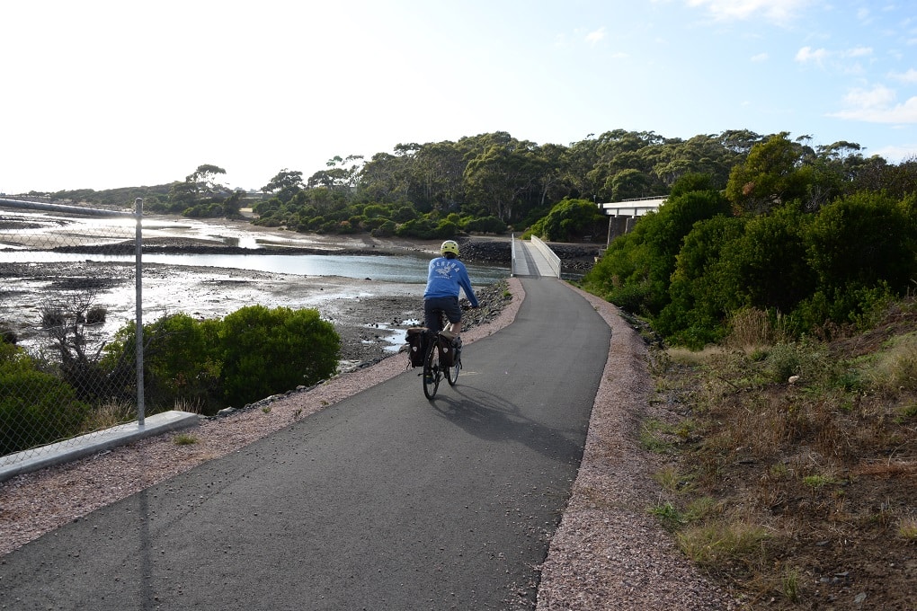 A man on a bicycle rides down an asphalt path towards a low bridge spanning a river and heading into bushland, with coastline visible tot he left.