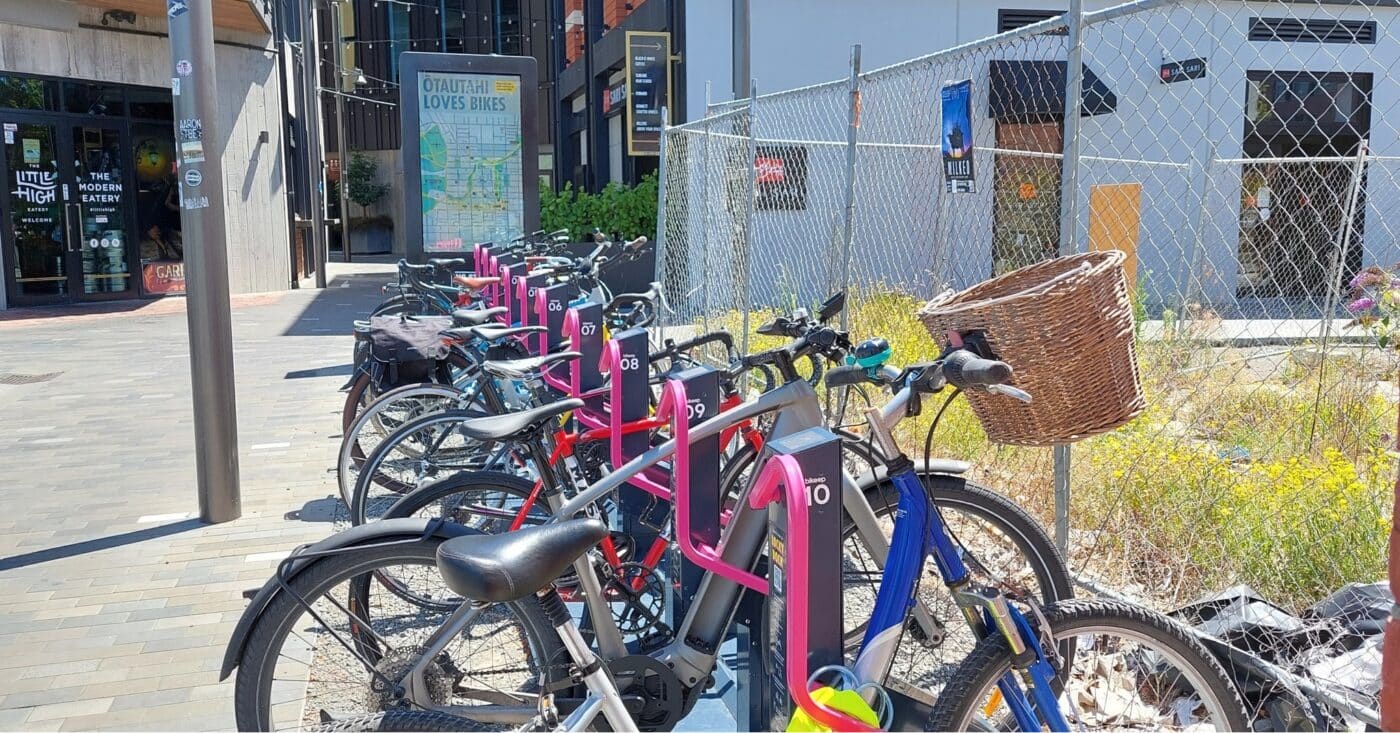 Row of electronic bike parking docks with bikes parked under hot pink steel arms with sign board in the background.