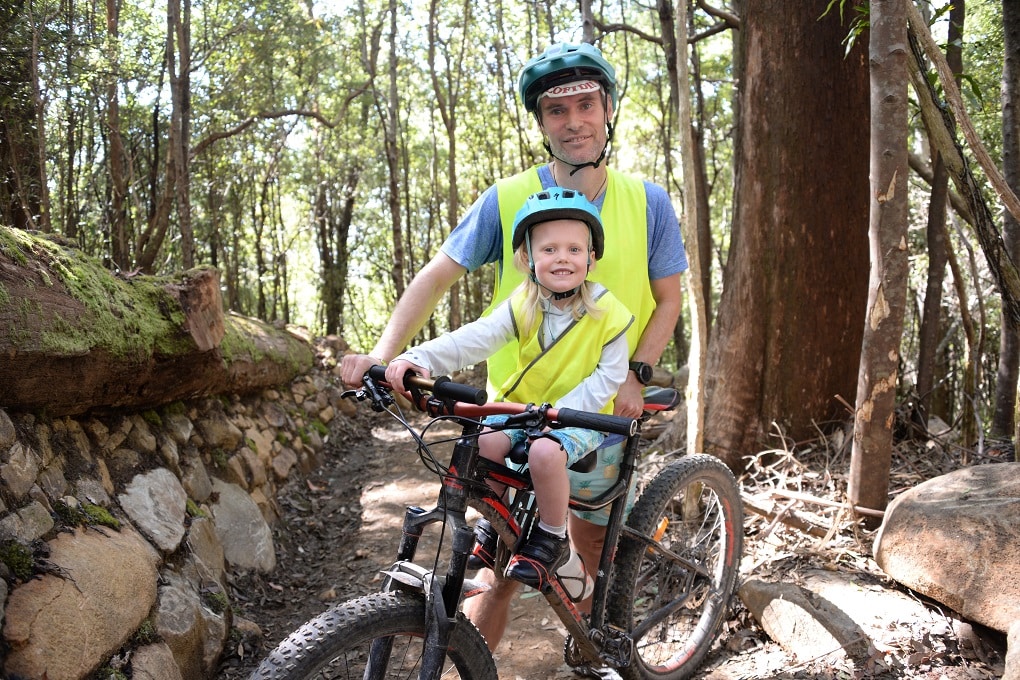 A man holds a mountain bike while a young girl sits in the shotgun saddle on the crossbar, both wearing blue helmets and yellow safety vests, surrounded by forest.
