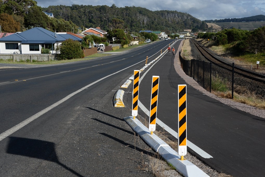 A road with  a section delineated by concrete kerbing and yellow and black striped bollards runs next to rail lines with two people riding in the section.