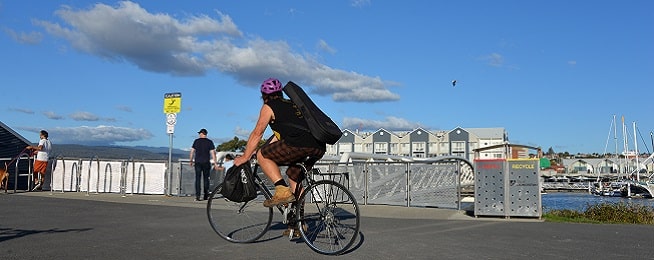 A man rides a bicycle along the Riverbend path with a guitar slung over his back and bag on his handlebars, looking towards the Seaport building on the other side of the river.