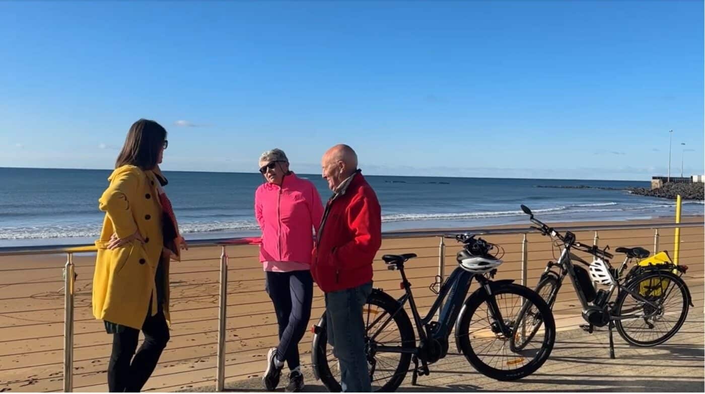 Mayor Teeny Brumby wearing a yellow overcoat leans against a fence overlooking the beach, talking to rider advocators Alison Horch and Keith Price whose bikes are leaning agains the fence.