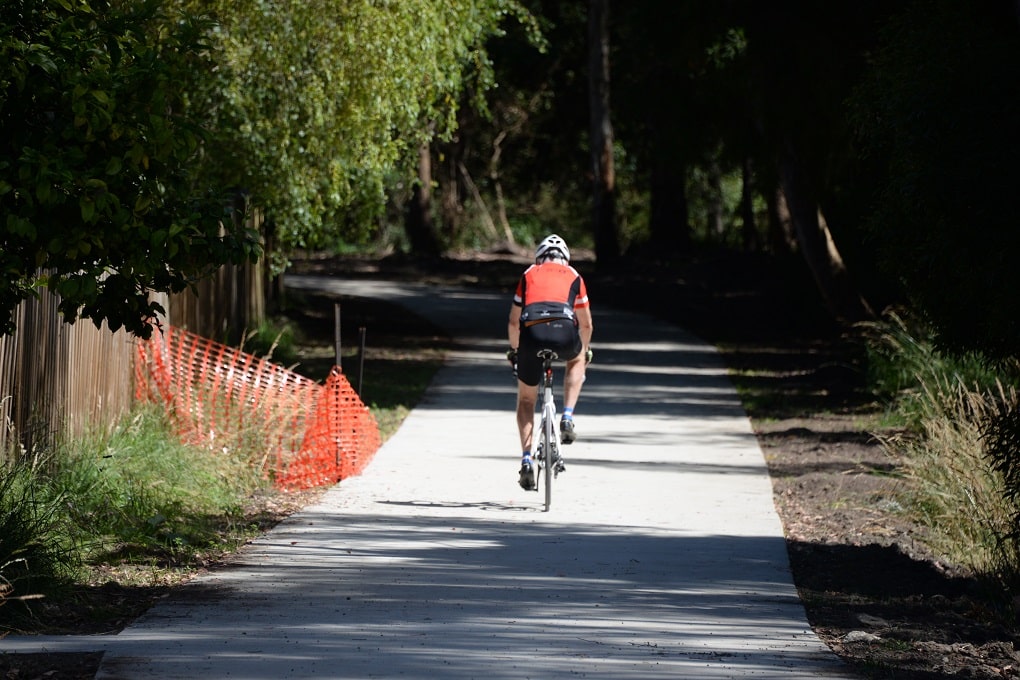 Man wearing red and black lycra kit rides along a side concrete path through bushland.