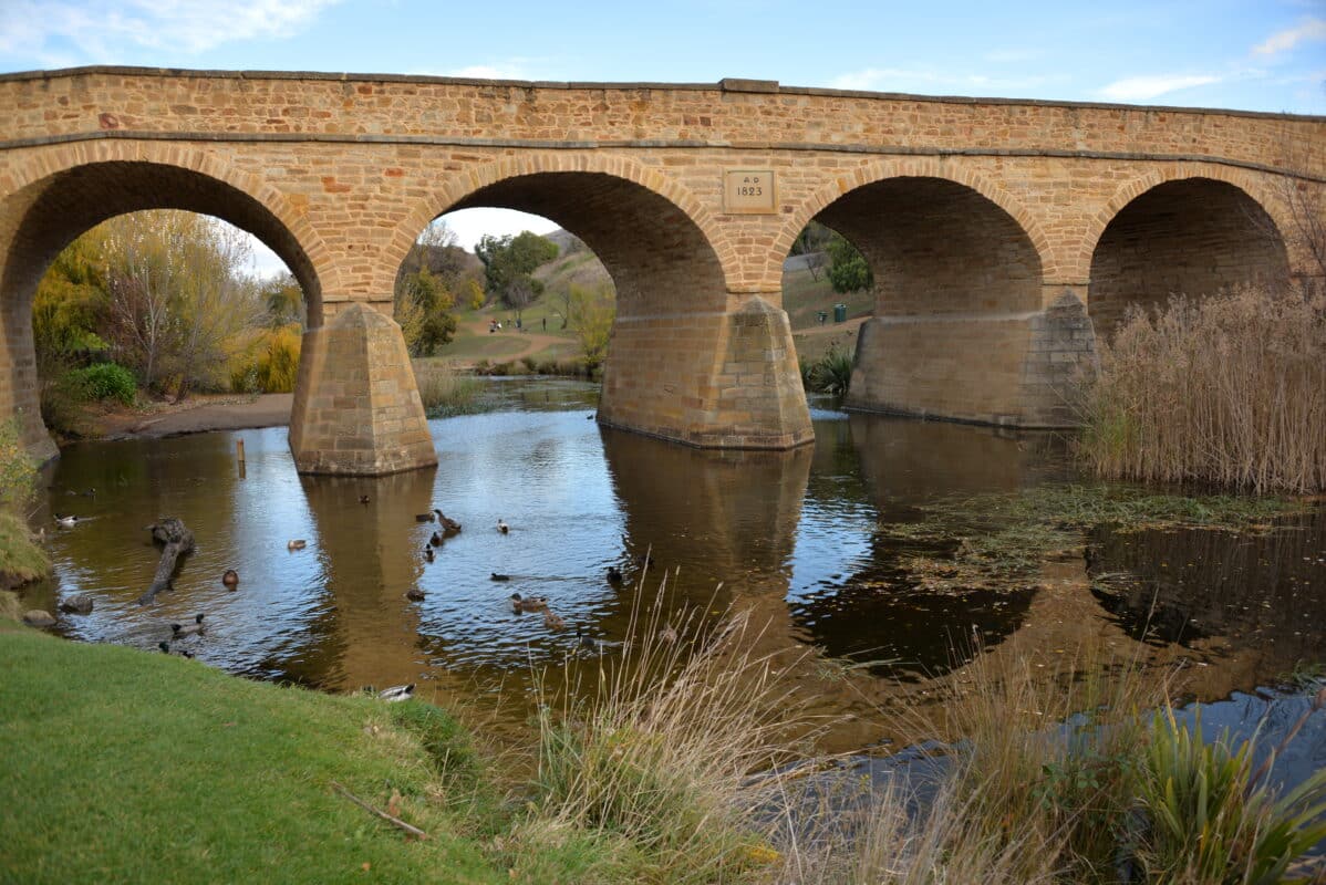 An old sandstone bridge with archers spanning a low river and grassy banks and blue sky.