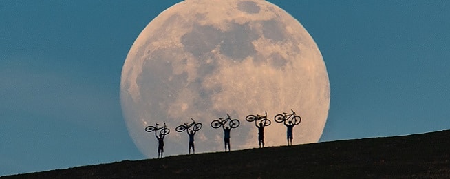 A rising moon in the background provides the backdrop for five people holding their bikes over the heads, in silhouette against the moon.