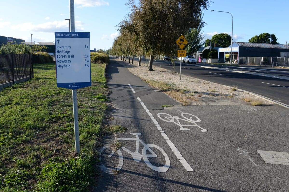 A blue and white sign next to a shared path with bicycle symbols separated from a road by a row of trees.