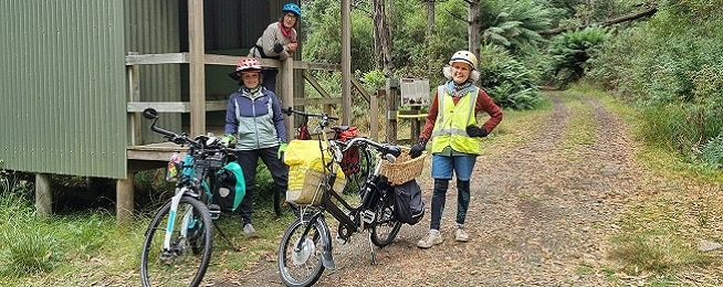 Three older women stand on a gravel path surrounded by forest with their bikes, with one woman leaning over a rail in a shelter.