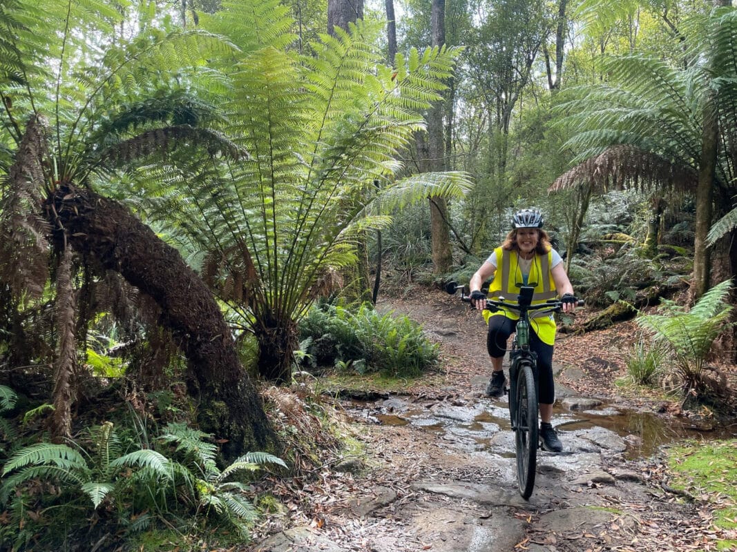 Redheaded woman wearing a yellow high vis vest smiles as she rides through a puddle on a gravel path flanked by tree ferns and forest.