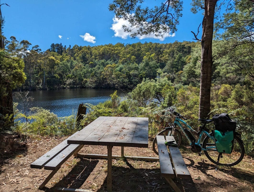 A bike leans against a wooden picnic table with a blue sky and white fluffy clouds over  a river edged by forest.
