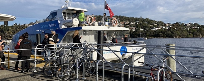 The blue and white Derwent Ferry docked at Bellerive with people boarding and three bikes locked to hoops in the foreground.