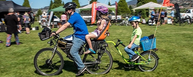 Man rides an electric bicycle on green grass with young child in seat on the pack rack and another child attached on a bicycle to the main bicycle.