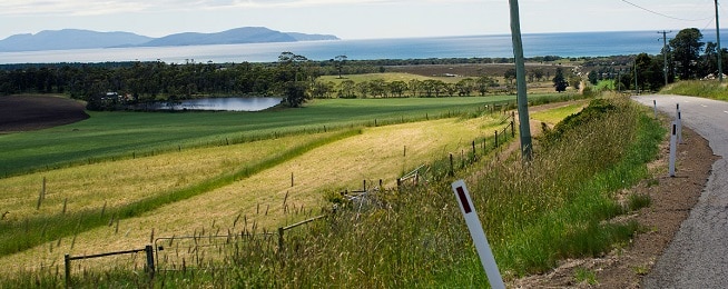 a winding rural road to the right with green fields and small dam in the foreground and coast with water and distant hills in the background.
