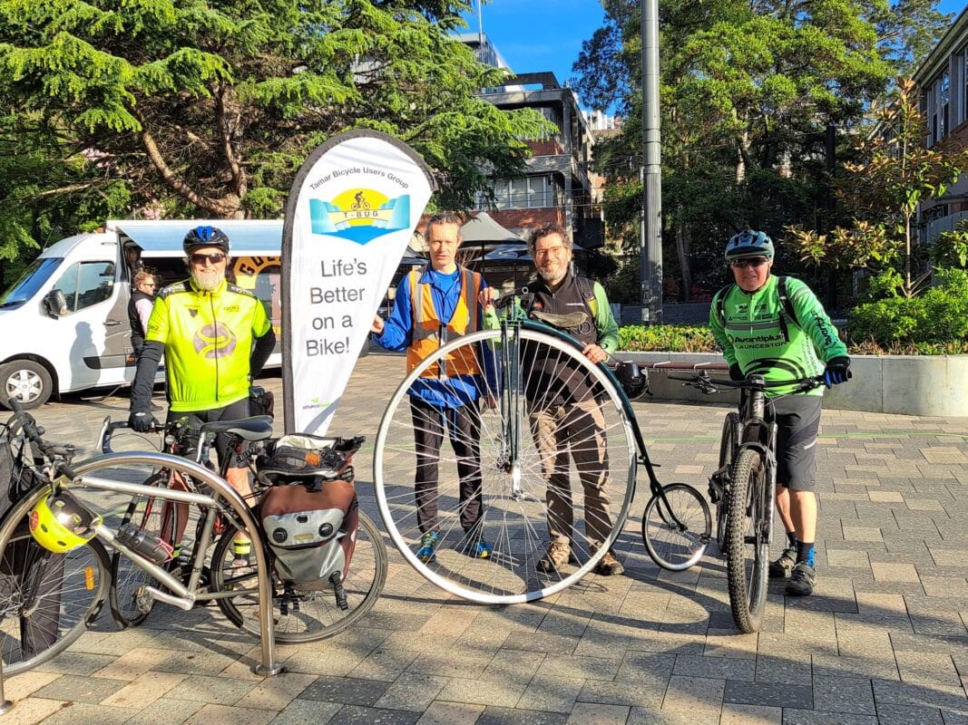 A group of men with bicycles, including a penny farthing, stand in an open space next to to a white flag with the TBUG logo on it.