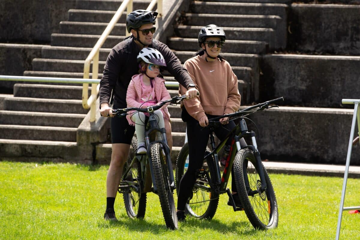 An adult women sits on a bike next to an adult man on a bike with a young child sittng in front of him on the bike crossbar.