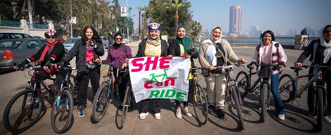 A group of women wearing hijabs stand iin a line with their bicycle with the middle women holding a sign saying She Can Ride.