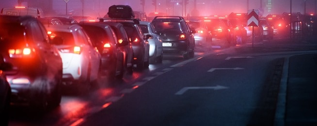 Cars at a standstill at night on a road with all the lights on and smoggy/foggy conditions.