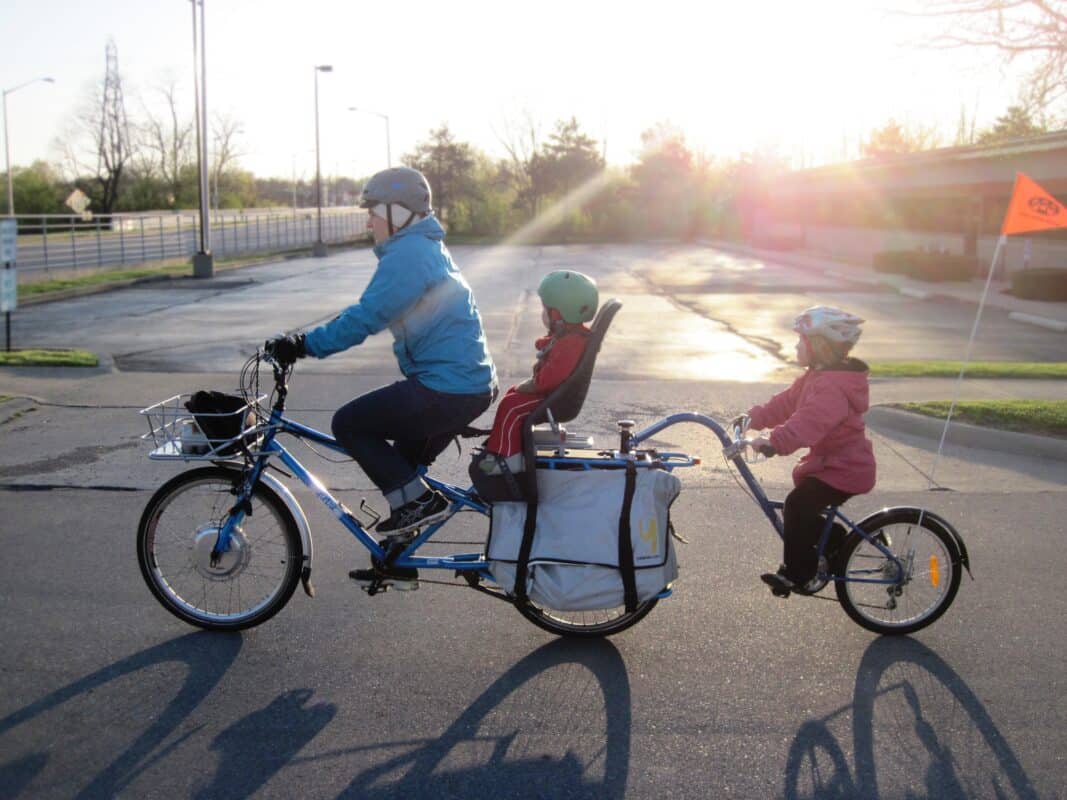 An adult rider on an electric Yuba cargo bike with a toddler in a seat behind, loaded panniers and a child attached to a bike at the back.
