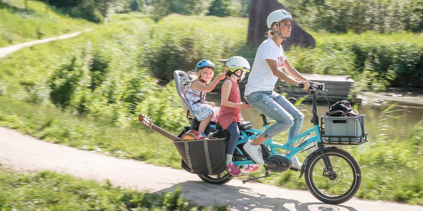 A woman wearing tshirt and jeans rides a blue Tern cargo bike along a path through a park with two young girls seated on the back and a guitar sticking out of a pannier. 