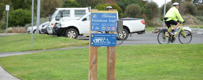 A blue and yellow sign for the Clarence Foreshore Trail is in the centre of the image, with a man wearing a bright yellow jacket riding in the background.