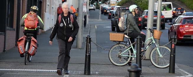 Three women at an intersection in Hobart. One is walking towards the camera, one is side on astride her bike waiting to cross the road and the other is riding away from the camera wearing a backpack with two panniers on the back of the bike.