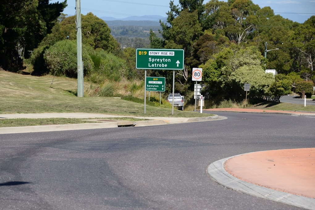 Image shows roundabout exit with large green and white traffic sign saying "Stony Rise Rd" and an arrow with Spreyton and Latrobe written below.