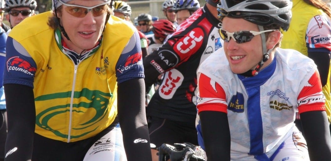 Two young men in cycling gear talk to each other with other cyclists in the background.