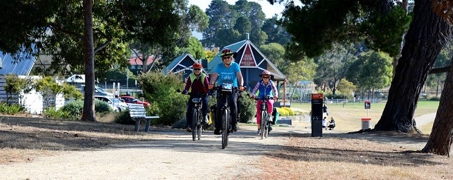 Three women ride in an arrow formation towards the camera on a gravel path with trees and a building behind them.