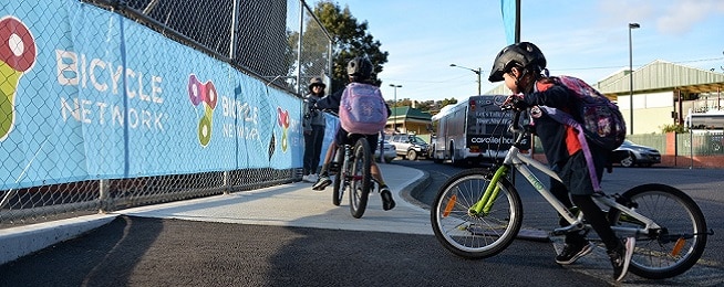 Two young girls ride up onto a footpath with their mother waiting for them alongside a fence that has bright blue Bicycle Nework signage tied to it .