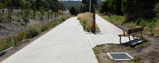 A wide concrete path running alongside a road separated by trees and fence on the left of the image, with another concrete path forking off to the right of the image with a wooden bench seat and sign at the junction of the two paths.