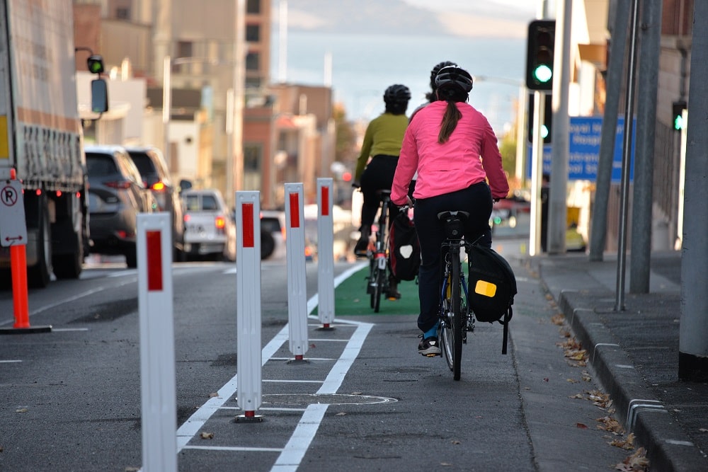 Three riders heading towards Liverpool St ride on a painted bike lane with white bollards in the road to their left, with the last rider wearing a bright pink jacket.