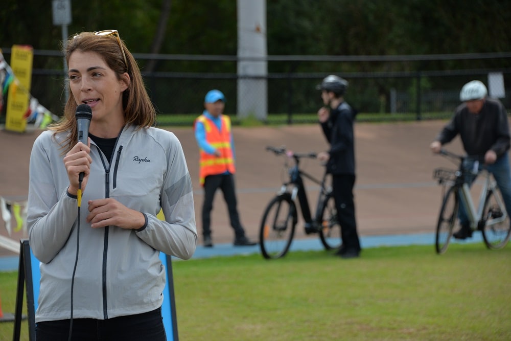 A woman holds a microphone with people with bikes in the background and a man in a high vis vest directing them.