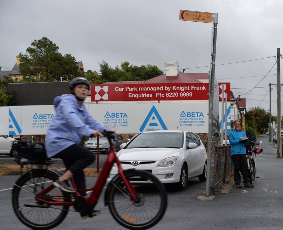 A woman riding a red step through bike with violet jacket is slightly blurred in the foreground with woman wearing blue jacket leans against a sign directing people to the Rivulet Path, smiling at the camera. 