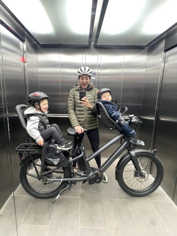 Woman takes photo in a lift of herself holding a bicycle with a young boy sitting on the front and young boy sitting o the back of the bike.