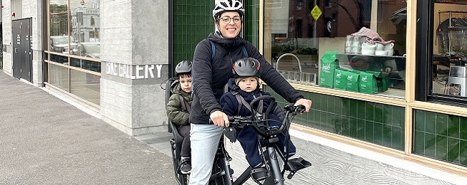 Woman rides a bicycle with a young boy at the front and back of the bicycle.
