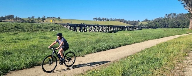 Man rides along gravel path with old wooden bridge in the background.