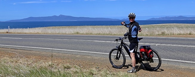 Man stands next to road astride a bicycle with dry grass on either side and the sea and mountain in the distance.