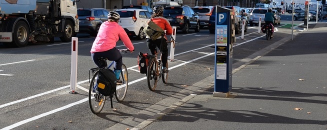 Three people ride on a painted bike lane with white bollards in the road to their left, with a blue parking meter on the footpath to their right and traffic in the road alongside the lane.