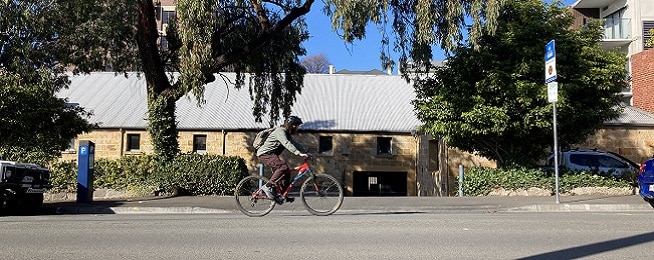 A man rides a mountain bike along Collins Street, with a sandstone building in the background and blue sky.