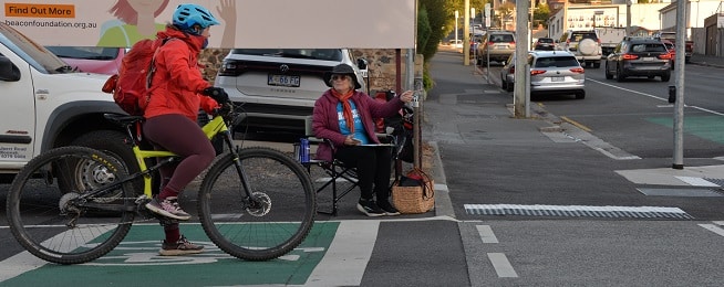 A woman on a bike stopped at an intersection talks to a woman sitting in a folding chair with clipboard on her her lap.
