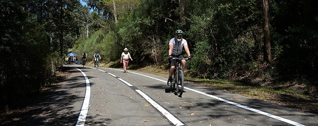 A man rides a bicycle on a new asphalt path with white line marking flanked by bush. Behind him a man walks and more people riding are in the distance.